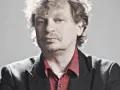 Marek Zieliński, aktor, w czarnej marynarce i czerwonej koszuli, z kręconymi włosami.