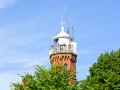 Widok na ceglaną latarnię morską w Ustce z tarasem widokowym, na którym stoją ludzie. Wokół zielone drzewa i błękitne niebo.