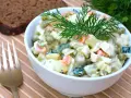 Ein Schüsselchen Olivier Salat, garniert mit Dill, neben einer Scheibe dunklem Brot und einer Gabel.