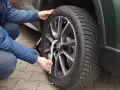 Osoba w niebieskiej kurtce dokonuje wymiany koła w samochodzie, używając klucza do odkręcenia śrub.