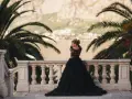 Elegancka kobieta w czarnej sukni z falbanami pozuje na tle palmy i morza. To jej wyjątkowe wesele na czarno.