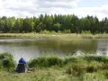 Mężczyzna w niebieskiej bluzie siedzi na krześle nad jeziorem, łowiąc ryby. W tle las i trzciny. Idealne miejsce na relaks, jak w **kiekrz**.
