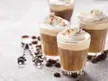 Tre bicchieri di deliziosa crema al caffè, guarniti con panna montata e cacao. Chicchi di caffè e cucchiaini decorati completano la scena.