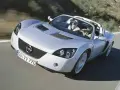 Srebrny Opel Speedster z dwoma pasażerami pędzi po krętej drodze.