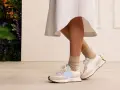 Elegancka sukienka i sportowe buty tworzą stylowy kontrast. Nogi w beżowych skarpetkach i białych sneakersach z błękitnym logo.