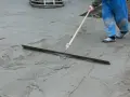 Pracownik w niebieskich ogrodniczkach wygładza świeży beton długą pacą. Trwa wygrzewanie posadzki.