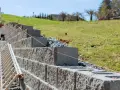 Budowa muru oporowego na skarpie. Betonowe bloki układane są warstwami, a różowa nić wyznacza poziom. Obok stoi drabina.