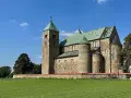 Kamienny zamek w Tumie z zielonym dachem, otoczony murem i zieloną łąką, pod błękitnym niebem.