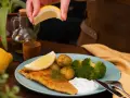 En hand pressar citron över en tallrik med panerad torsk, potatis, broccoli och en klick sås.