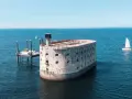 Fort Boyard, potężna twierdza na morzu, otoczona błękitną wodą. Obok platforma wiertnicza i żaglówka.