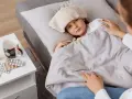 Chora dziewczynka z okładem na głowie leży pod kocem. Obok leki i kubek. Mama czuwa, zapewniając opiekę na dziecko.