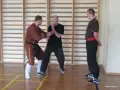 Trzech mężczyzn w sali gimnastycznej, dwóch w strojach do sztuk walki, ćwiczy ruchy.