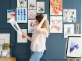 Mujer joven sonríe mientras revisa un dibujo de perro, rodeada de acuarelas de flores, aves y un flamenco. Un espacio inspirador para estudiar dibujo.