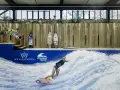 Frau surft auf einer künstlichen Welle in Berlin. Mehrere Surfbretter stehen bereit.