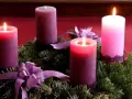 Drei brennende Kerzen auf einem Adventskranz aus Tannengrün mit lila Schleifen. Die dritte Kerze leuchtet hell.