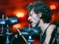 Intensywna fotografia koncertowa: perkusista z kręconymi włosami i zarostem, z zamkniętymi oczami, uderza w bębny.
