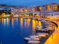 Port na Minorce o zmierzchu, z łodziami zacumowanymi w zatoce i oświetlonymi budynkami.