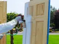 Persona con guantes y traje protector aplica laca blanca a una puerta de madera con pistola pulverizadora. Proceso de como lacar puertas.