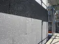 Gros plan sur des blocs de polystyrène gris, certains recouverts d'une couche noire, en cours d'installation pour une isolation par l'extérieur. Échafaudages visibles à droite.