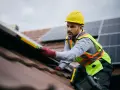 Instalator w żółtym kasku i kamizelce bezpieczeństwa montuje panele fotowoltaiczne na dachu. To dowód, że fotowoltaika jest bezpieczna, gdy montują ją profesjonaliści.