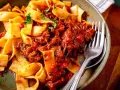 Breite Bandnudeln mit herzhaftem Ragu-Sauce, garniert mit frischer Petersilie. Ein Gabel liegt bereit.