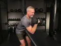 Man med grått hår och skägg utför preacher curls med en hantel i ett gym.
