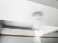 Detector de humo blanco en techo, emitiendo luz roja y humo. Cumpliendo la normativa de detectores de humo para seguridad.
