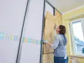 Frau baut eine Trockenbauwand, setzt Dämmmaterial in Metallständer ein.