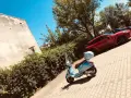 Błękitny skuter czeka na przejażdżkę. W tle czerwony samochód i zielone drzewa. Idealne miejsce na parking moto.