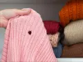 Różowy sweter z dziurą, odkryty podczas przeglądania stosu ubrań w szafie. Widać inne swetry w różnych kolorach.