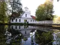 Biały kościół na wodzie w Zwierzyńcu, z odbiciem w tafli jeziora i mostkiem.