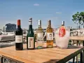 Pięć butelek wina, w tym jedno bordeaux, na drewnianym stole na tarasie z widokiem na miasto.