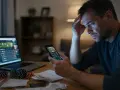Zmartwiony mężczyzna z telefonem i laptopem, na których widać strony bukmacherskie. Stosy żetonów i rachunków sugerują problem z hazardem w internecie.