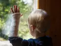 Kind mit Gips am Arm, das durch ein Fenster schaut. Ein gebrochenes Handgelenk bei einem Kind ist schmerzhaft, aber die bunte Gipsbinde macht es erträglicher.