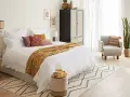 Habitación serena con cama blanca, manta ocre y cojines coloridos. Un armario de ratán y una silla acogedora complementan el estilo feng shui cama.