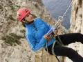 Junge Frau beim Abseilen an einer Felswand, Malta Klettern. Sie trägt einen roten Helm und eine blaue Jacke.