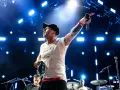 Wokalista z tatuażami i czapką na głowie prowadzi koncert One Republic, wskazując ręką w górę i trzymając tamburyn.