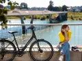 En kvinna i gula tröjan och jeans vilar vid en elcykel. Frågan "hur lång tid tar det att cykla 1 mil med elcykel" hänger i luften.