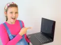 Uśmiechnięta dziewczynka w ogrodniczkach wskazuje na ekran laptopa, gdzie widać okienka z ikonami. Idealne miejsce na naukę angielskiego online dla dzieci.