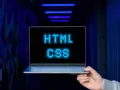 <p>Dłoń trzyma laptopa, na ekranie którego świeci napis "HTML CSS" w niebieskich pikselach. Tło jest ciemne, z niebieskimi neonami.</p>