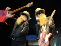 Dwóch muzyków z długimi, rudymi brodami, w kapeluszach i okularach, trzymających gitary. Jeden podnosi swoją gitarę nad głową, drugi pokazuje ją publiczności, tworząc wrażenie "over the under".