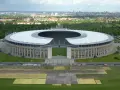 Das Olympiastadion Berlin mit seinem markanten Dach und der Skyline im Hintergrund. In der Ferne ragt der Fernsehturm, der wie ein glockenturm Berlin überragt.