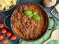 Herzhaftes Ragout Bolognese in einem Topf, garniert mit Basilikum. Daneben frische Tomaten, Bandnudeln und Parmesan.