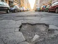 Un cratere nel marciapiede, con auto parcheggiate sullo sfondo. La gomma tagliata dal manto stradale crea un ostacolo pericoloso.
