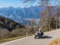 Motociclista su una delle **strade panoramiche del Lago di Garda**, con montagne imponenti e un villaggio in lontananza.
