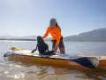 Frau richtet Sitz auf SUP-Board ein, das wie ein Kajak genutzt werden kann. Sonnenschein, Berge und See im Hintergrund.
