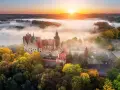 Zamek Książ otoczony jesiennymi drzewami, wyłaniający się z porannej mgły. Słońce wschodzi nad wzgórzami, malując niebo na pomarańczowo.