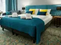 Przytulny pokój hotelowy z dużym łóżkiem, ozdobionym turkusową narzutą i żółto-niebieskimi poduszkami. Ręczniki z logo Stella Resort & Spa zachęcają do relaksu.