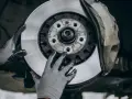 Mechanik w rękawiczkach wymienia zużyte tarcze hamulcowe. Widać nowy, lśniący element obok starego zacisku.
