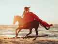 Kobieta w czerwonej sukni galopuje na koniu po plaży. Wiatr rozwiewa jej włosy i materiał sukni, tworząc dynamiczny obraz stadniny nad morzem.